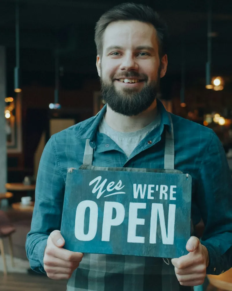 A café owner in an apron smiling and holding a “Yes, we’re open” sign